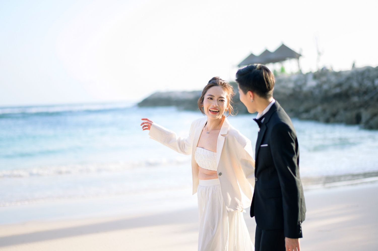 Couple in wedding attire on a sunny beach, bride in white dress pointing toward the ocean, groom in black tuxedo.