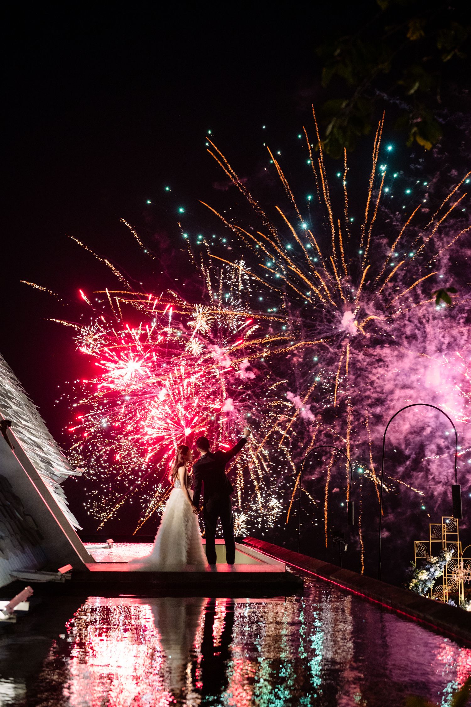 Bride and groom silhouetted beside a reflective pool as colorful red and pink fireworks light up the night sky at their wedding.
