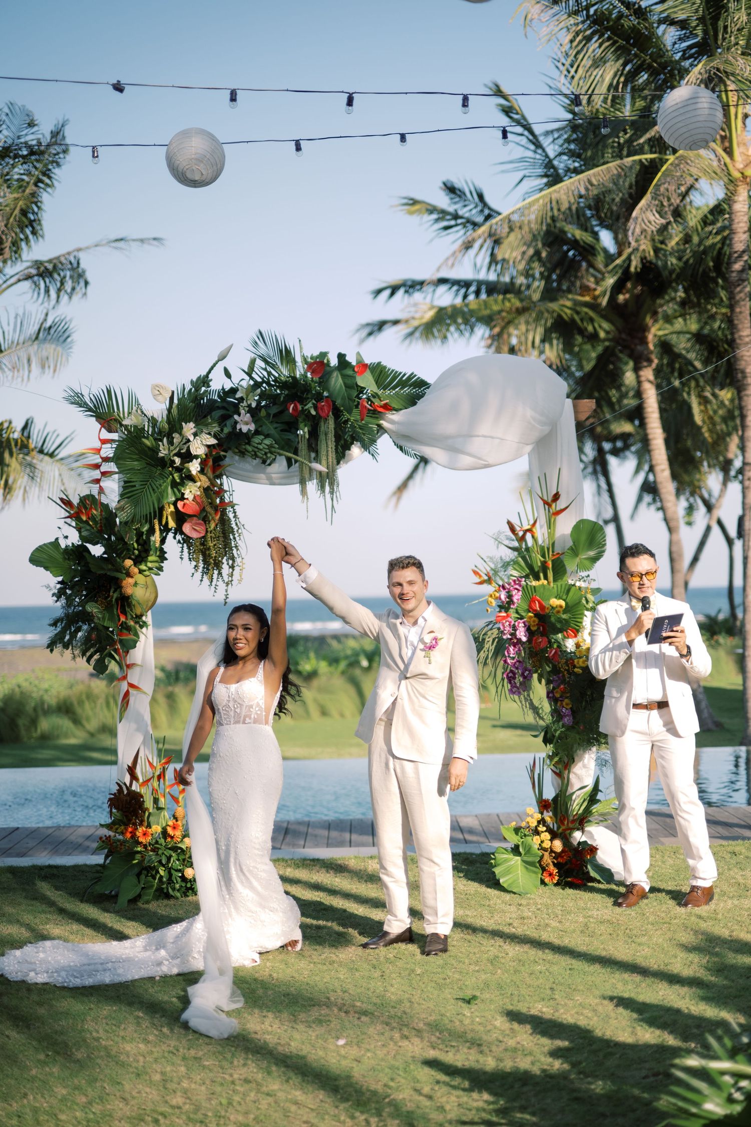 Couple celebrates at tropical beach wedding ceremony under floral arch with palm trees and ocean backdrop.