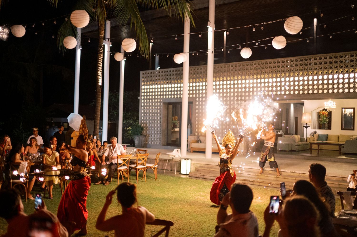 Fire dancers perform at an outdoor night event with white lantern decorations and an audience gathered on a tropical lawn.