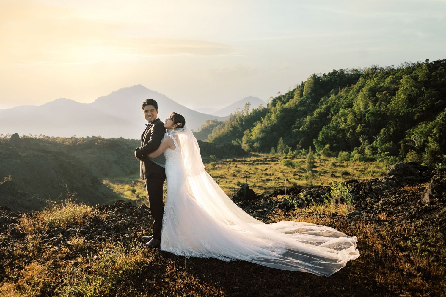 Couple in wedding attire posing on a scenic mountain overlook at sunset with lush green hills in the background.
