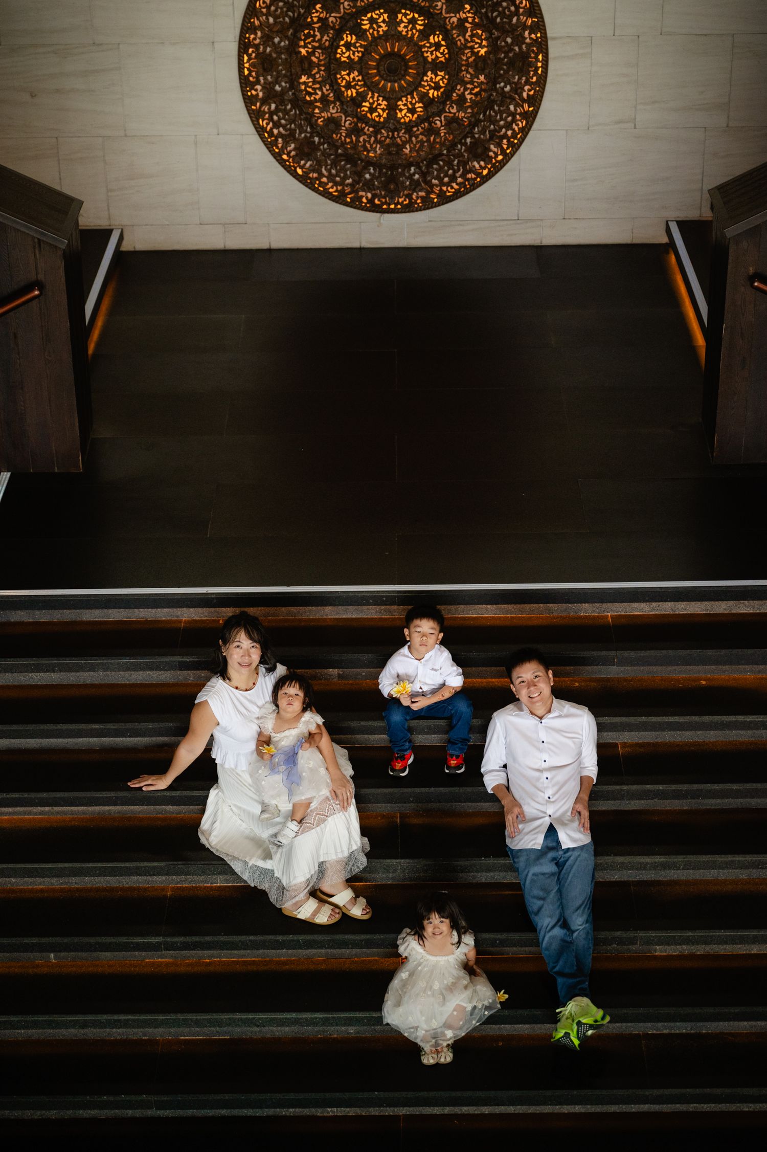 Family of four posing on grand staircase beneath ornate chandelier, dressed in white for a formal occasion.