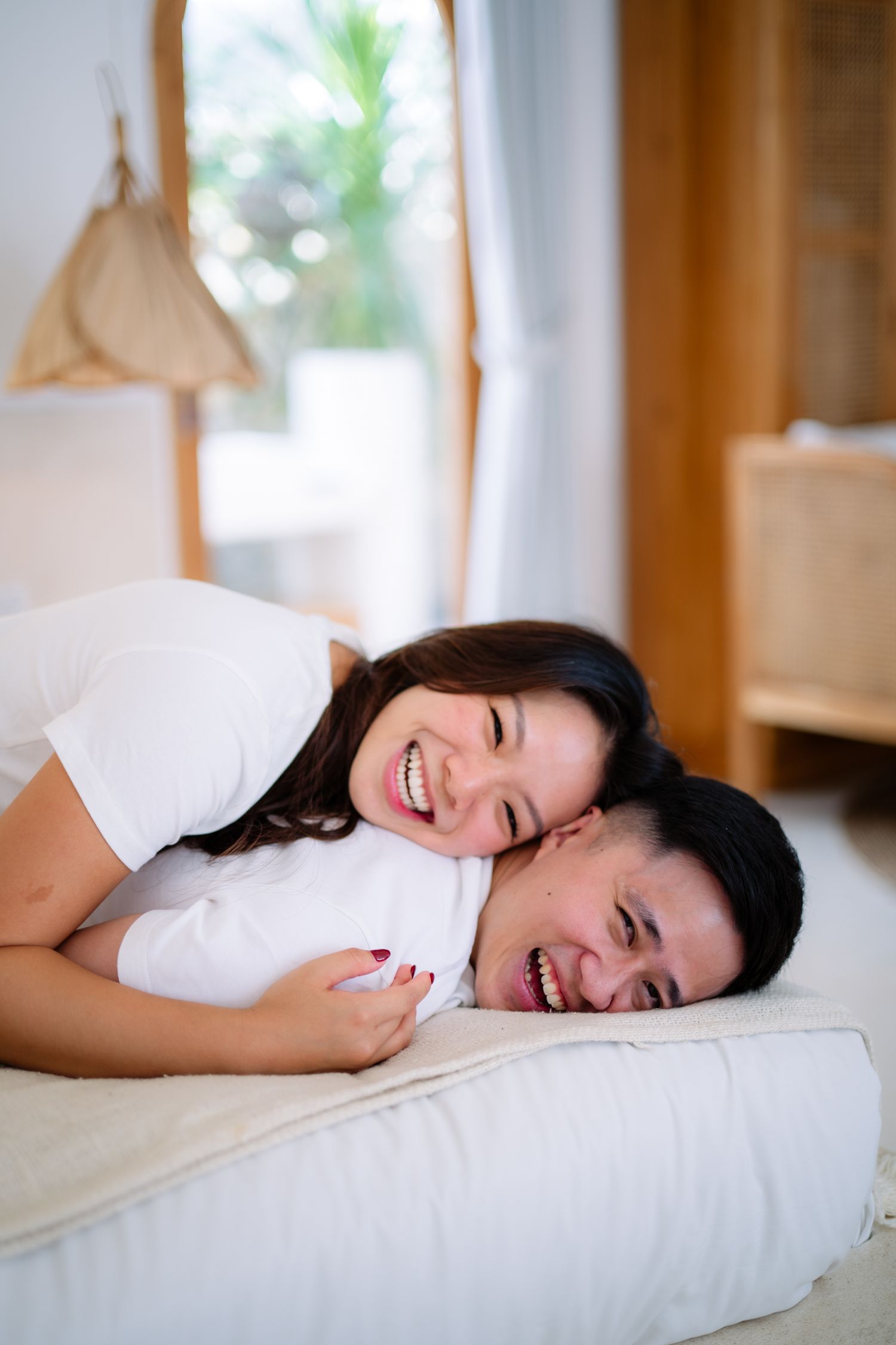 Happy Asian couple laughing and cuddling together on a white bed in a bright, cozy bedroom.