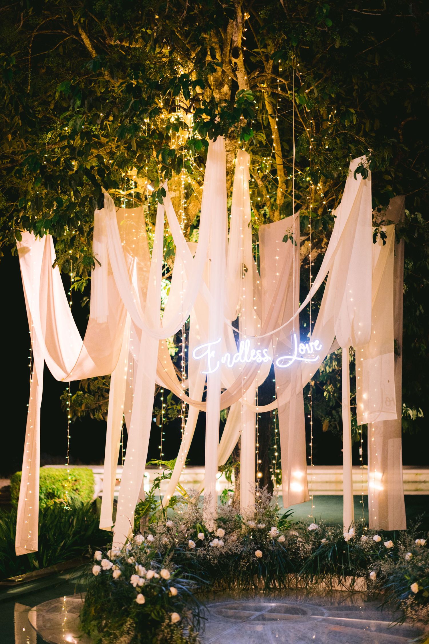 Elegant outdoor wedding backdrop with white flowing drapes, fairy lights, and neon 