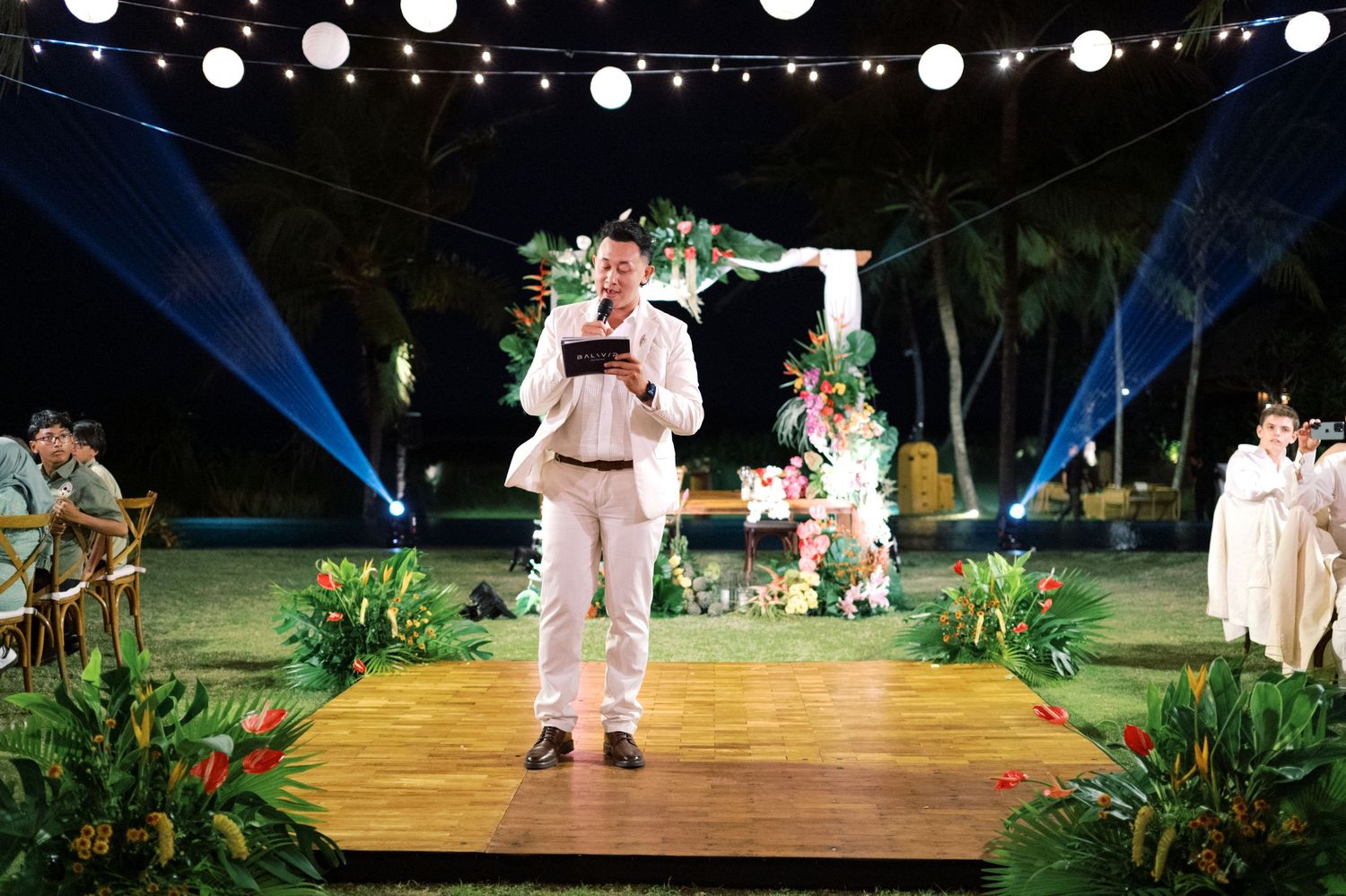 Man in white suit speaks at outdoor tropical wedding ceremony on wooden stage with lush greenery and lights.