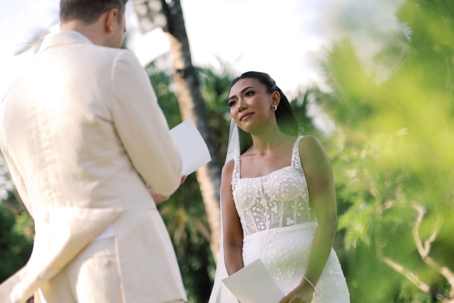 Bride in white lace gown listens emotionally as groom reads vows outdoors among palm trees.