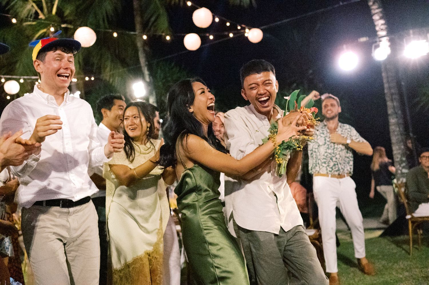 Wedding guests laugh and celebrate as someone catches the bridal bouquet at a tropical reception.