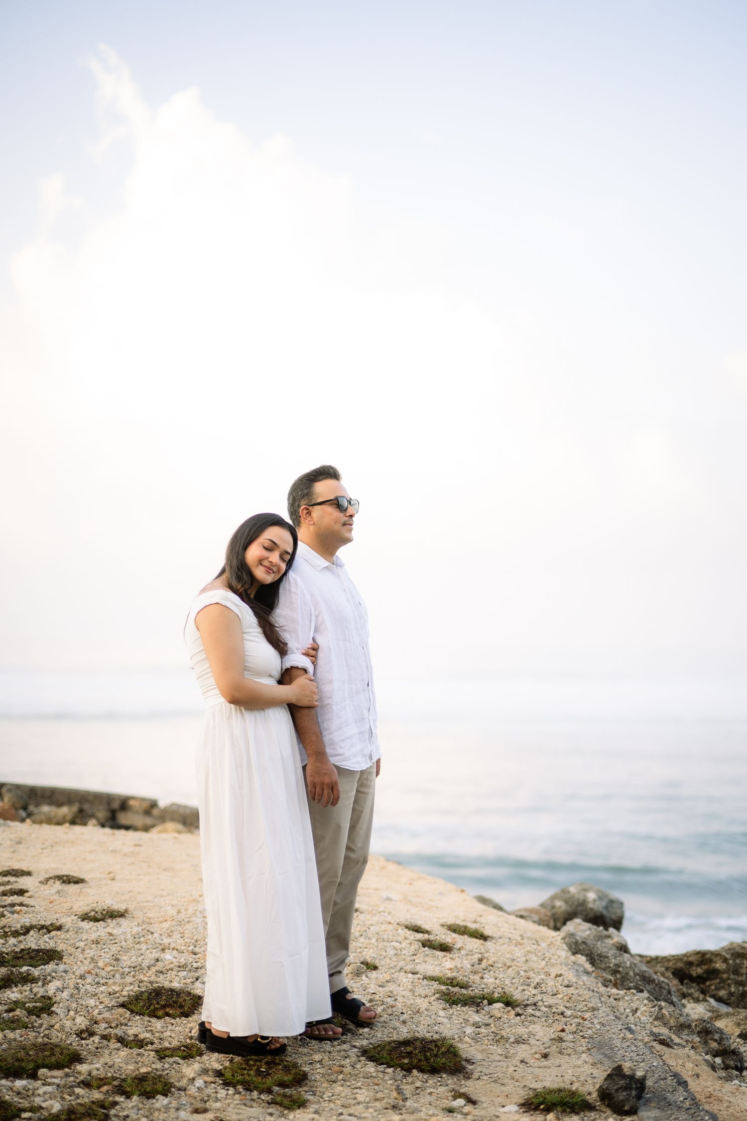 Couple in white outfits embracing on rocky beach shore with ocean backdrop at sunset.