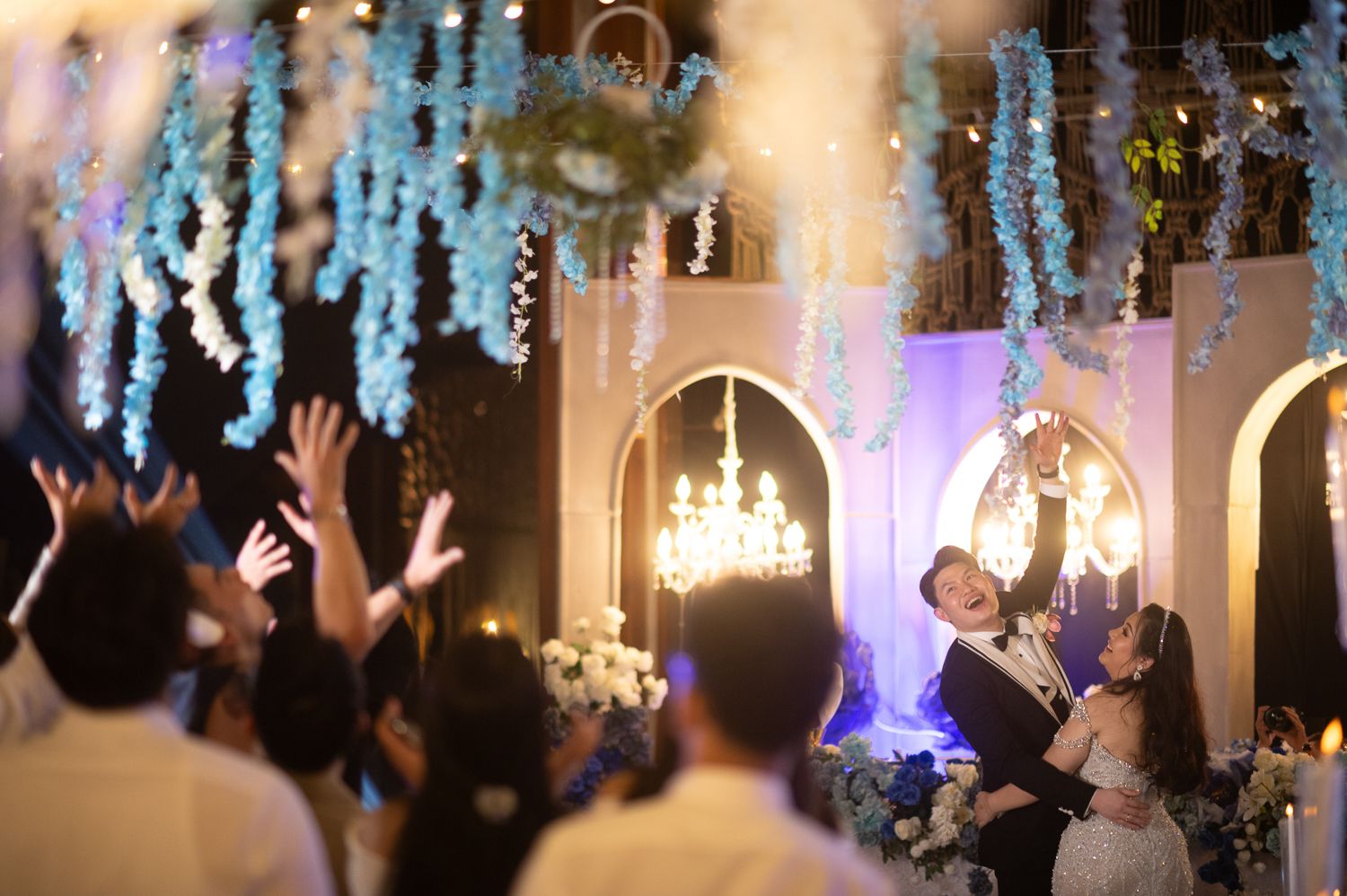 Elegant wedding reception with couple embracing on stage, guests raising hands, chandelier lighting, and blue floral decorations.