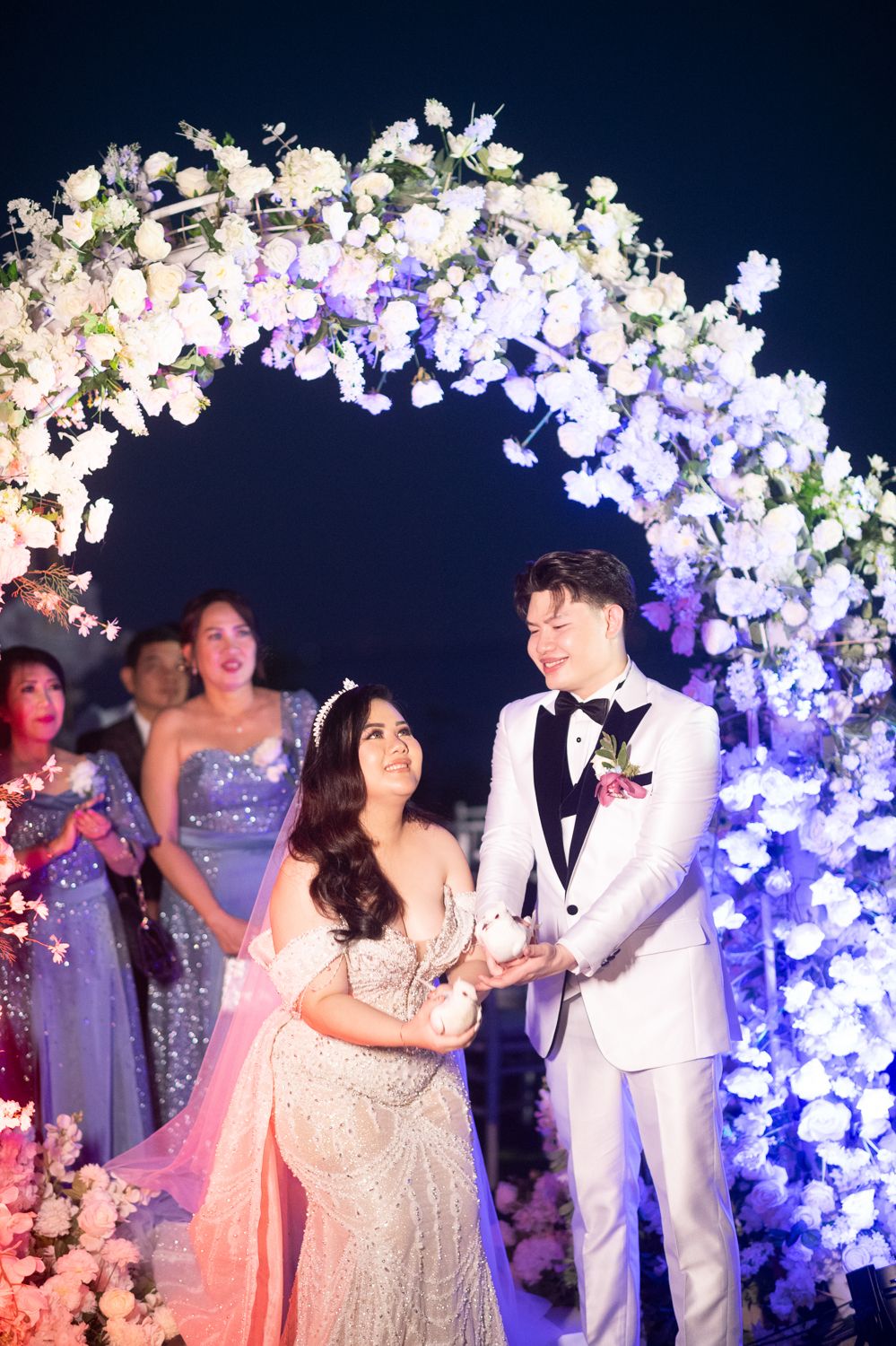 Couple smiling under illuminated floral arch at night wedding ceremony with guests in background.