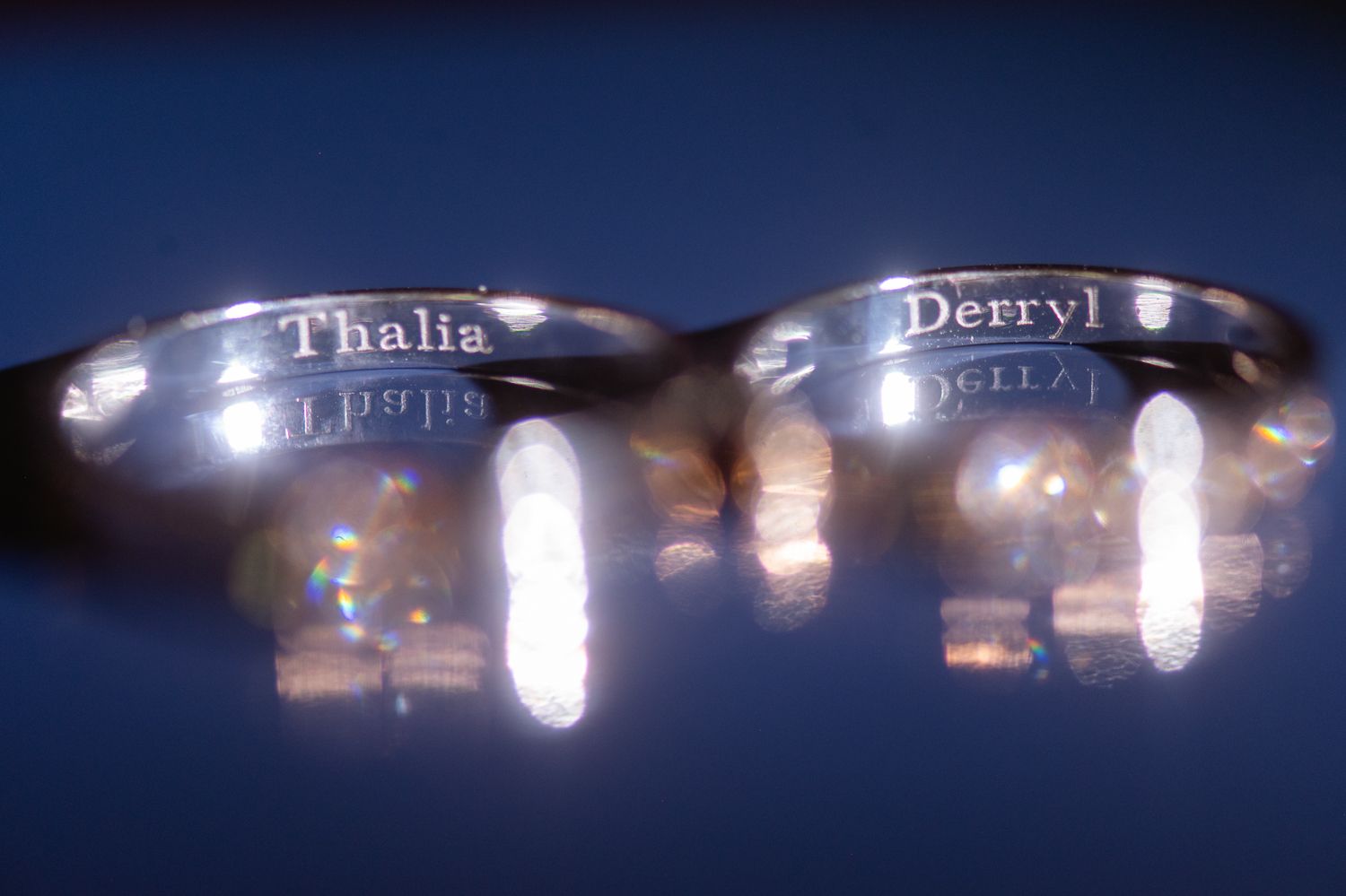 Two silver rings engraved with the names Thalia and Derryl, reflecting light on a dark surface.