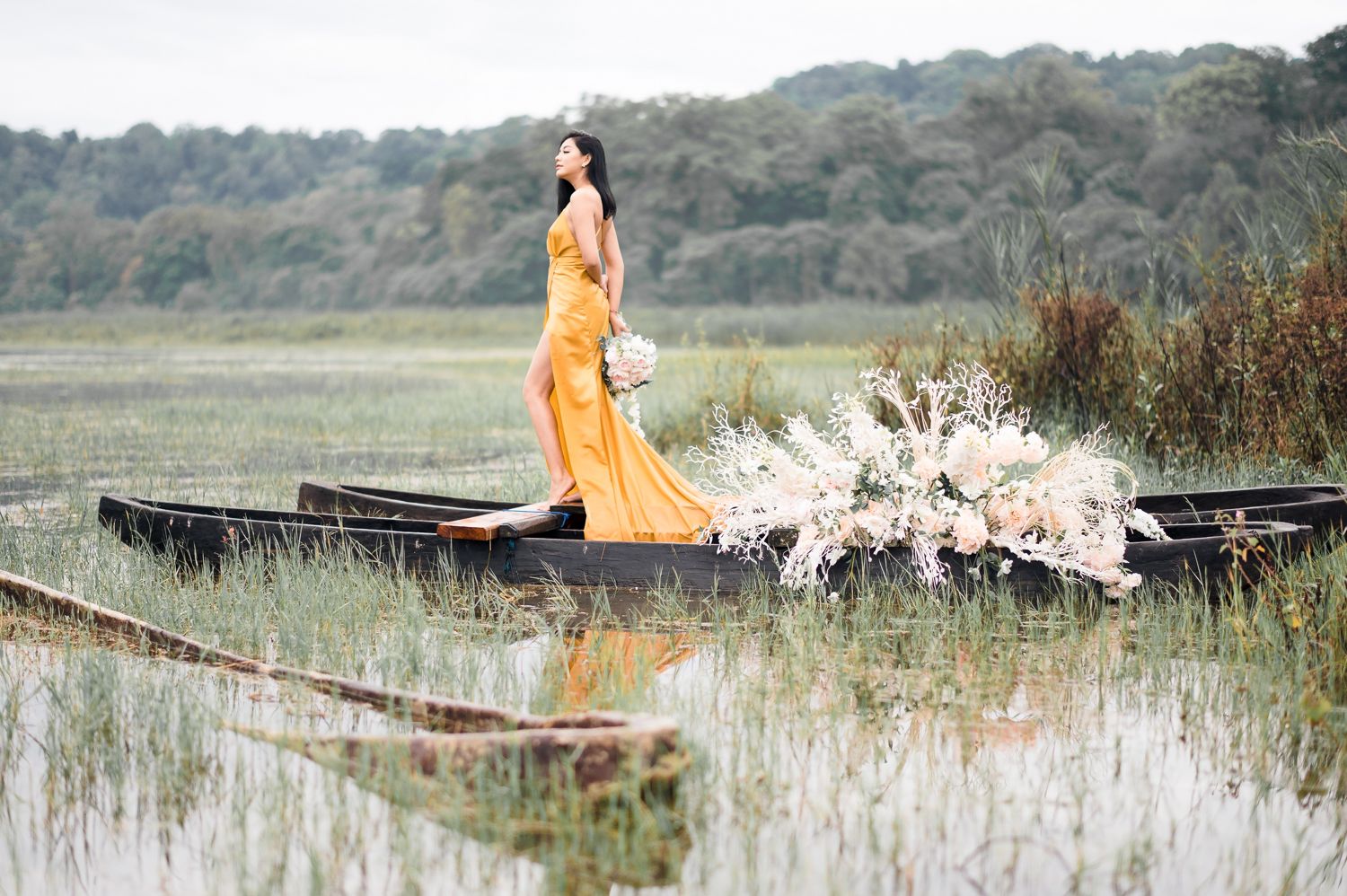 Woman in orange gown standing in a wooden boat surrounded by lush greenery and white floral arrangements on a calm lake.