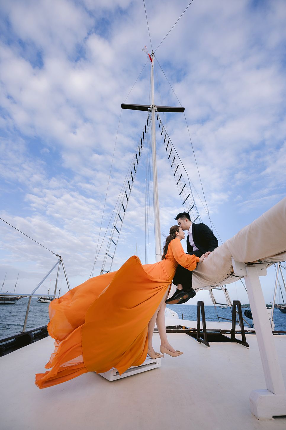 Couple in orange gown and black suit share romantic moment on yacht deck with blue sky backdrop.
