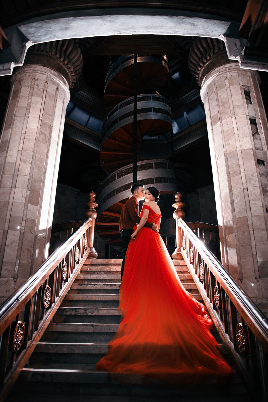 Couple kissing in red gown on grand staircase with spiral stairs and ornate columns in elegant venue.