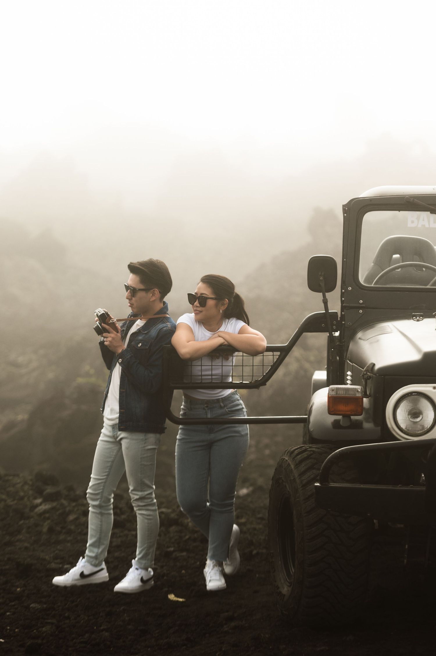 A couple in sunglasses poses beside a black jeep in misty volcanic terrain, man holding camera, woman leaning on vehicle.