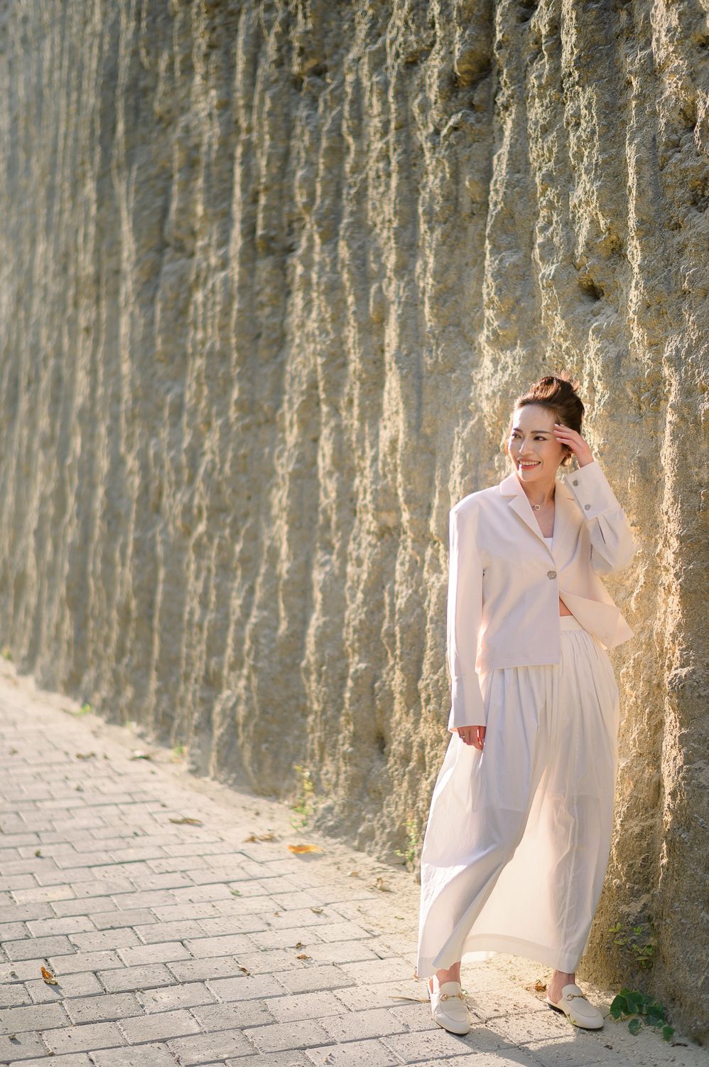 A smiling woman in a white outfit leans against a textured stone wall on a sunlit cobblestone path.