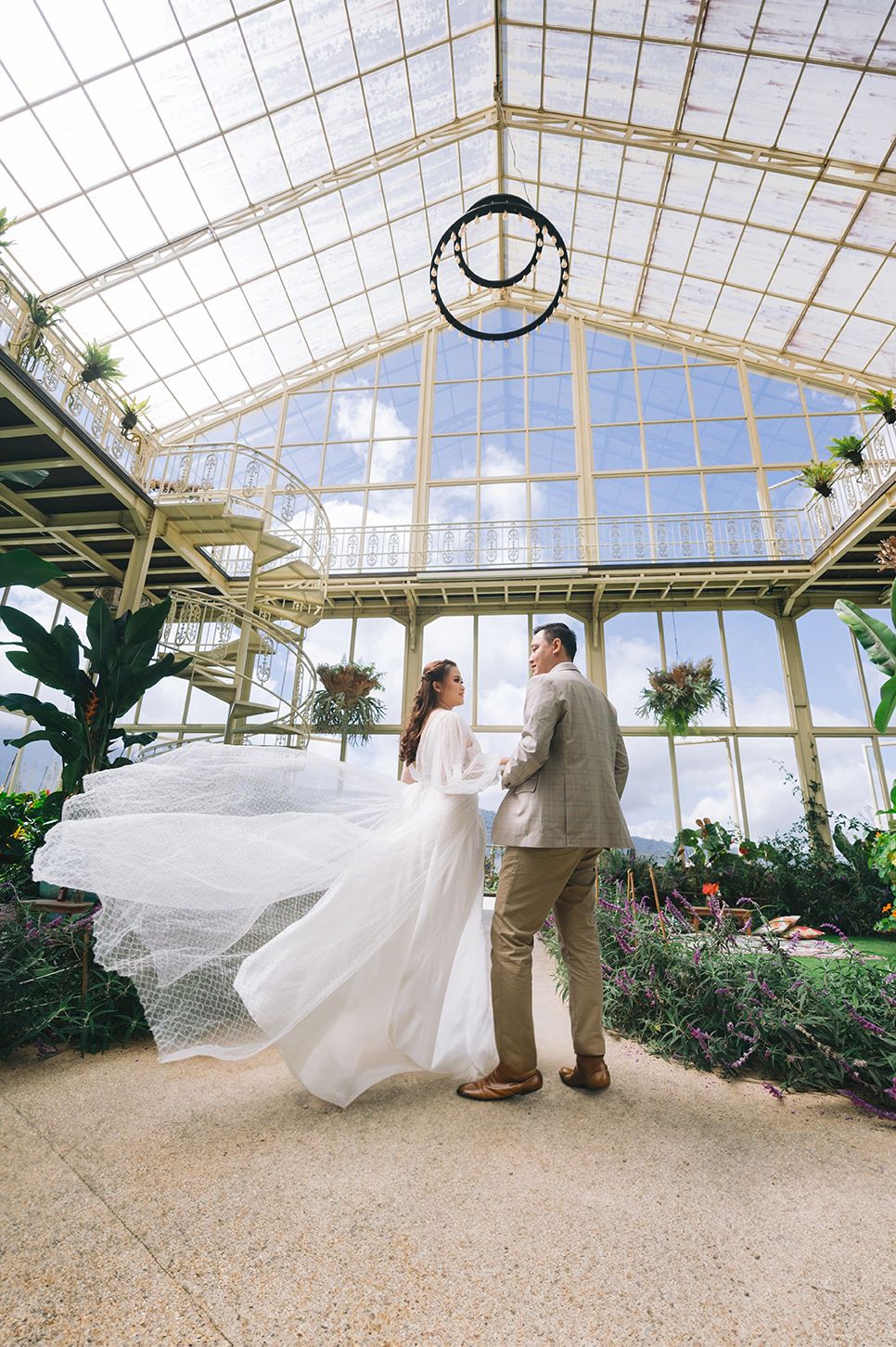 Couple in wedding attire posing inside a stunning glass greenhouse with lush greenery and flowing bridal veil.