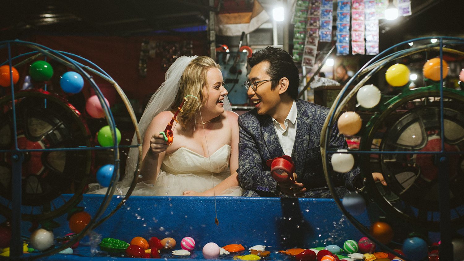 Bride and groom laughing together at a colorful carnival ball game during their fun wedding celebration.