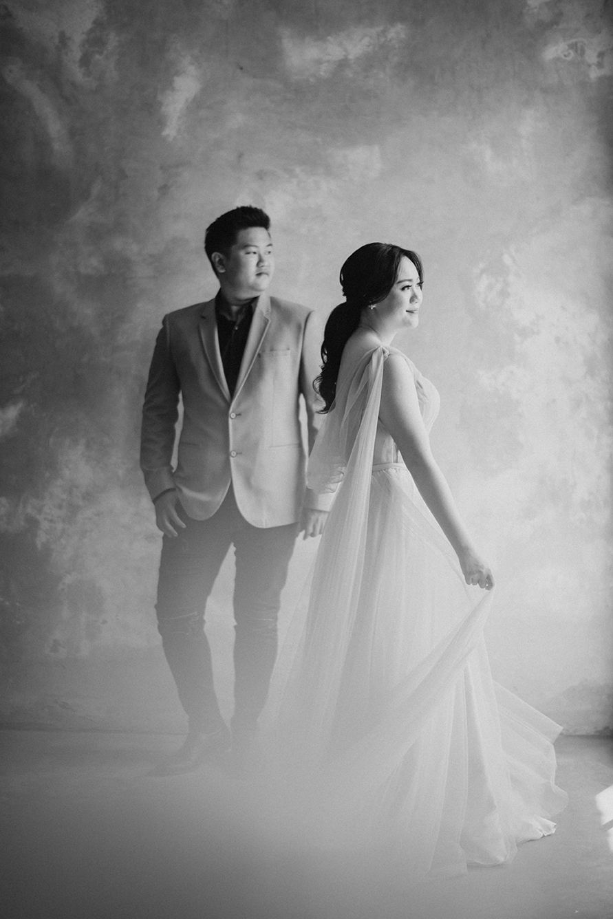 Black and white photo of an Asian couple in elegant wedding attire posing gracefully in a misty studio backdrop.