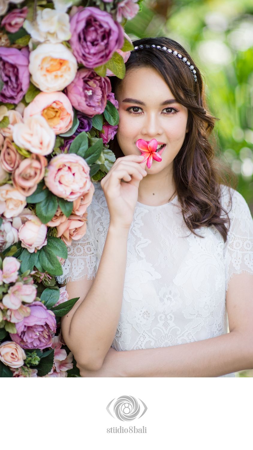 A bride in white lace dress holds a pink flower near a colorful floral backdrop of roses and peonies.