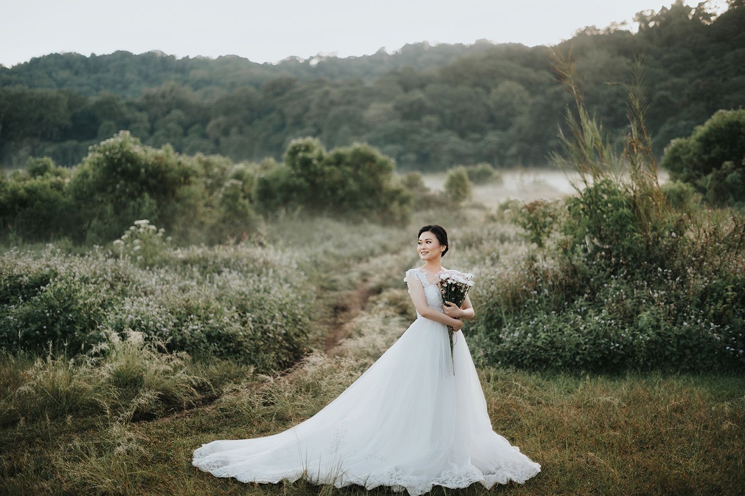 Bride in white ball gown holding bouquet stands in lush green meadow at golden hour.