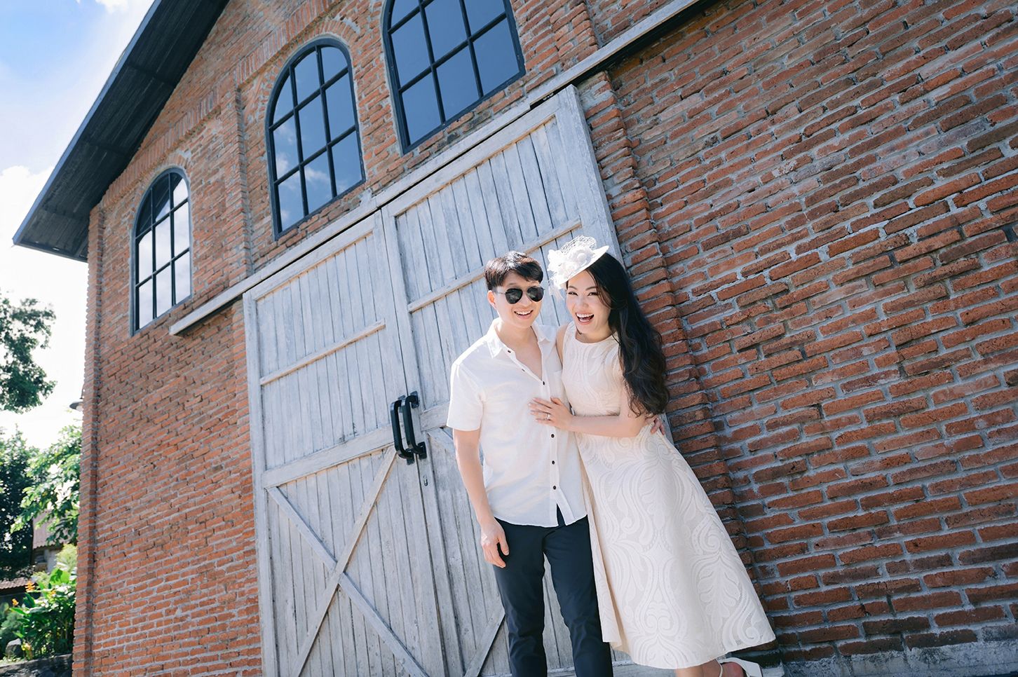 Stylish couple standing together in front of rustic brick building with large wooden doors on a sunny day.