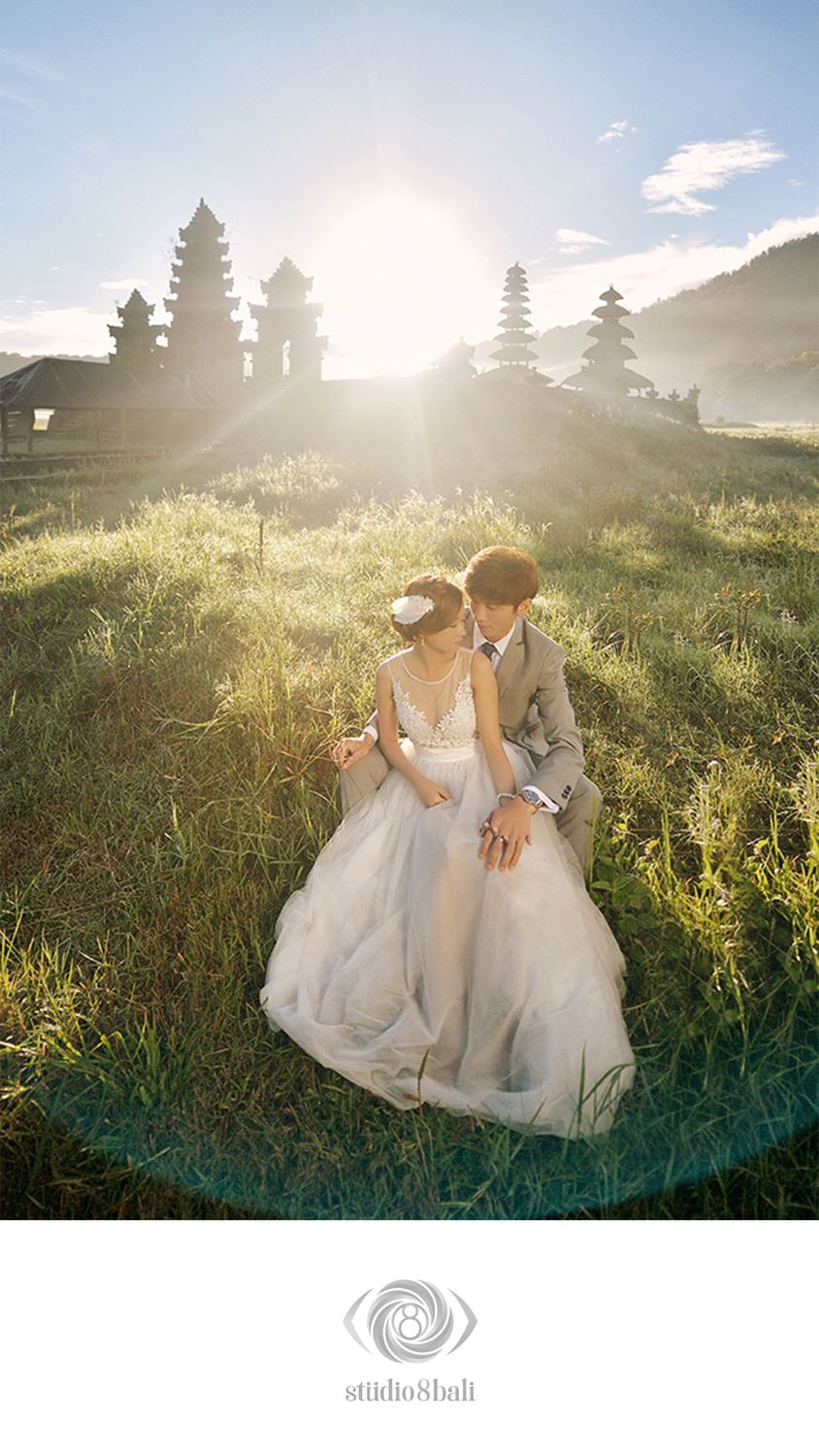 Couple in wedding attire sitting on grass with Balinese temple towers and golden sunlight in Bali.