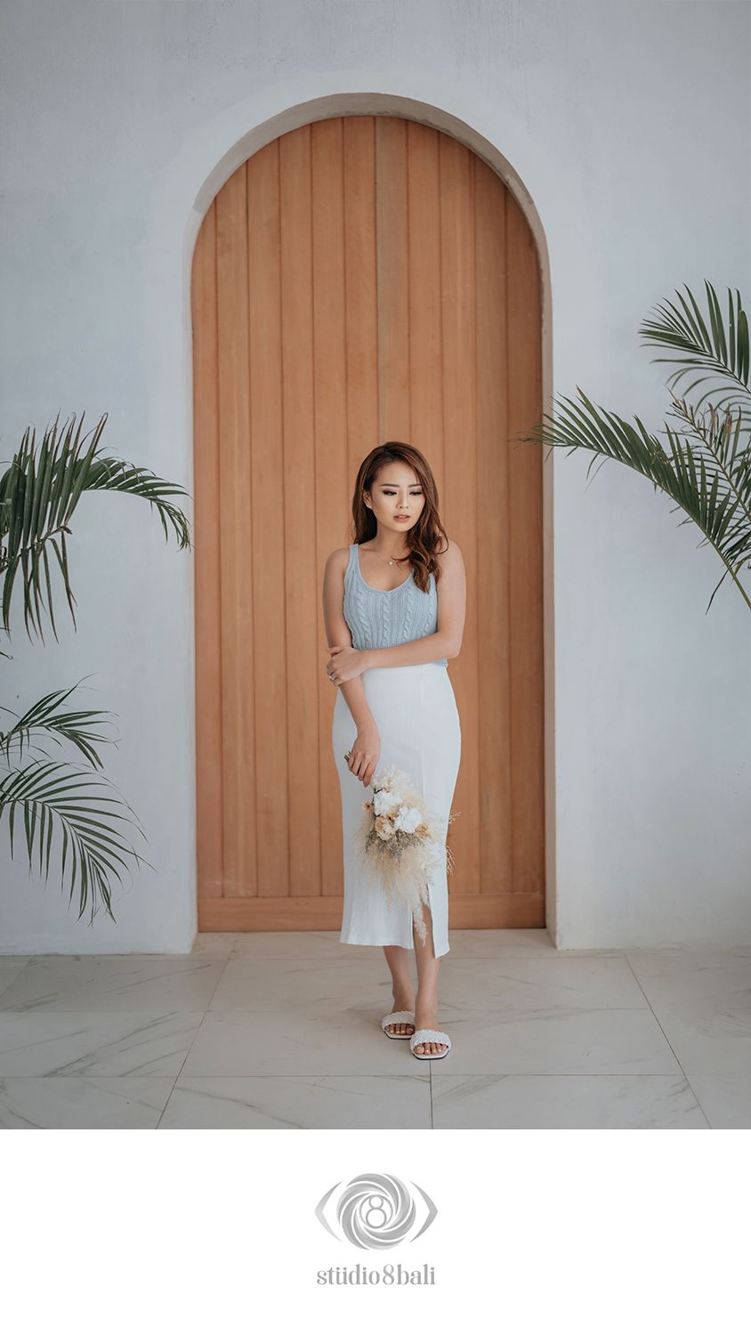 Woman in light blue top and white midi skirt holding bouquet, posing before arched wooden door with tropical plants.
