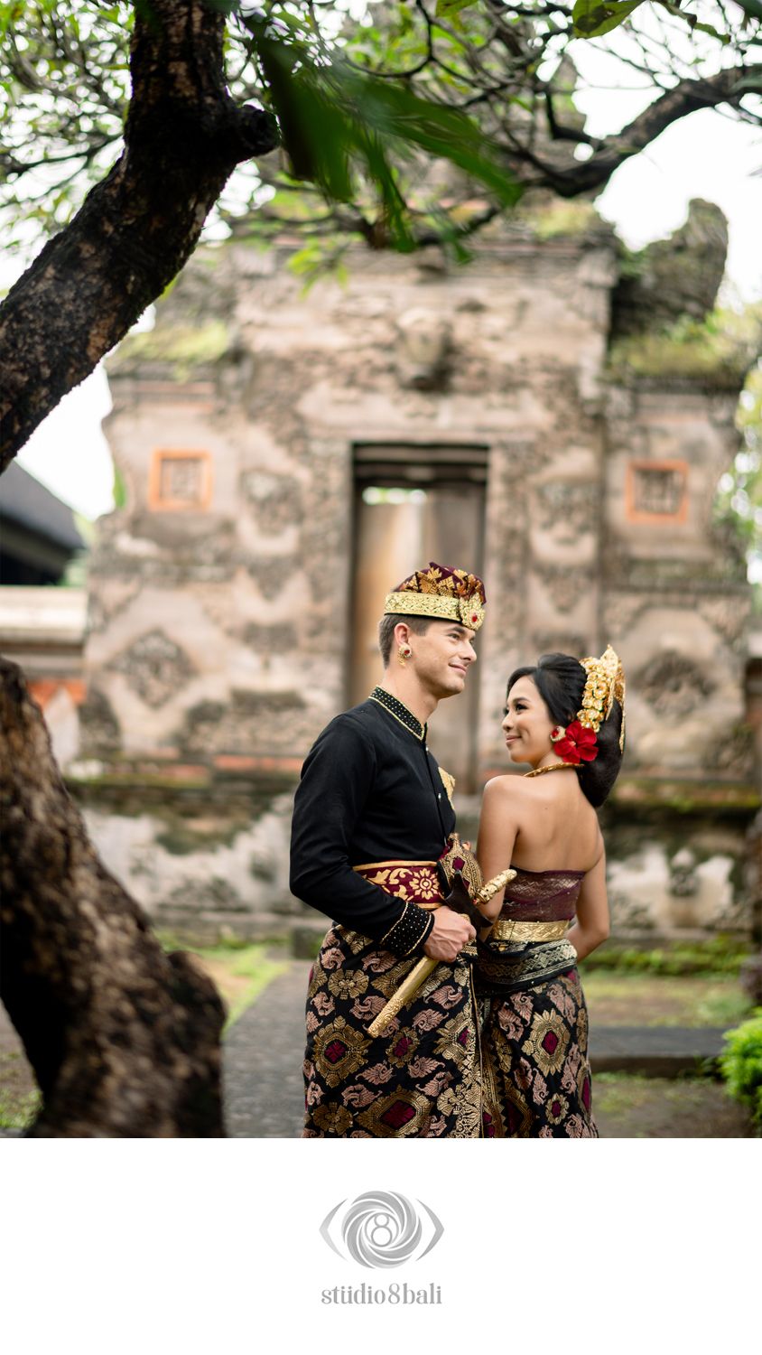 Balinese couple in traditional wedding attire posing before an ornate stone temple gate in Bali, Indonesia.