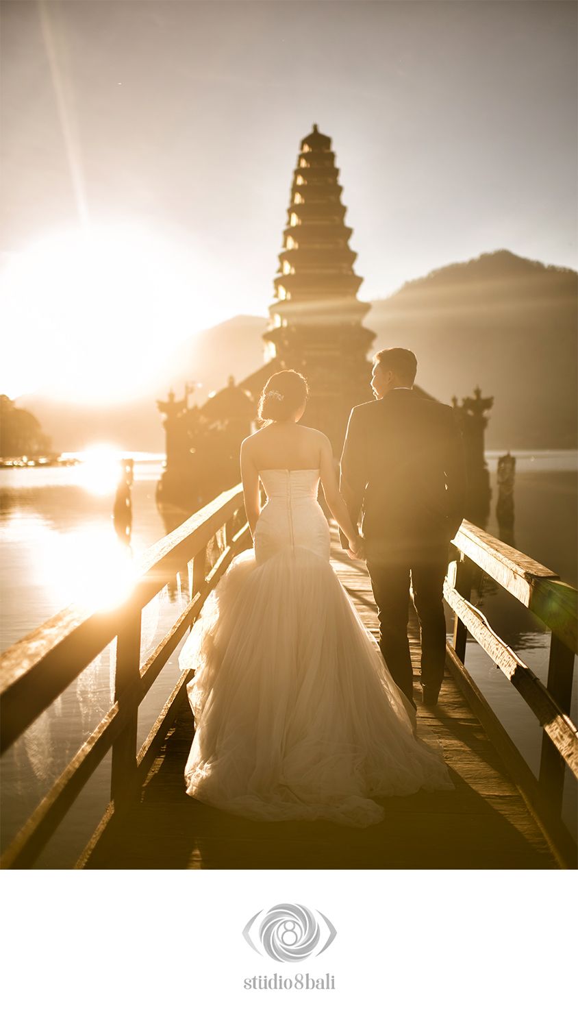 Couple in wedding attire walks toward a sunlit Balinese pagoda temple on a wooden bridge at golden hour in Bali.