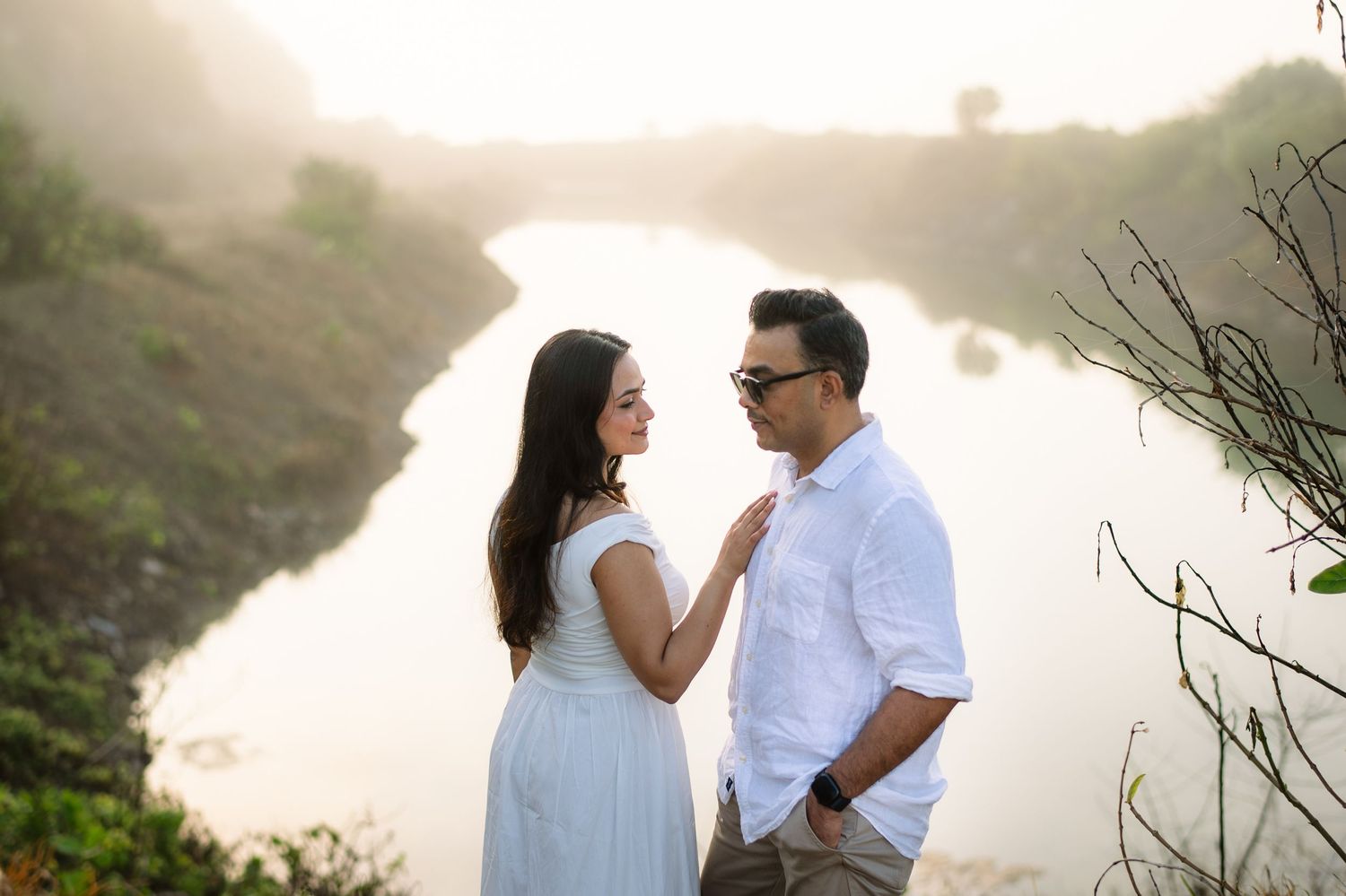 Couple sharing a romantic moment by a misty river at golden hour, woman in blue dress, man in white shirt.