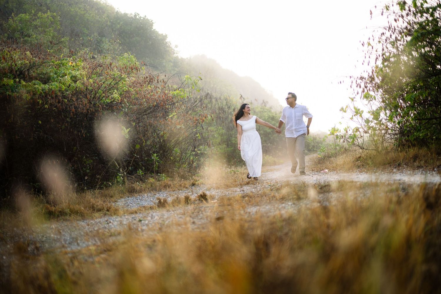 Couple holding hands walking on a scenic dirt path surrounded by lush greenery and golden grass in soft natural light.