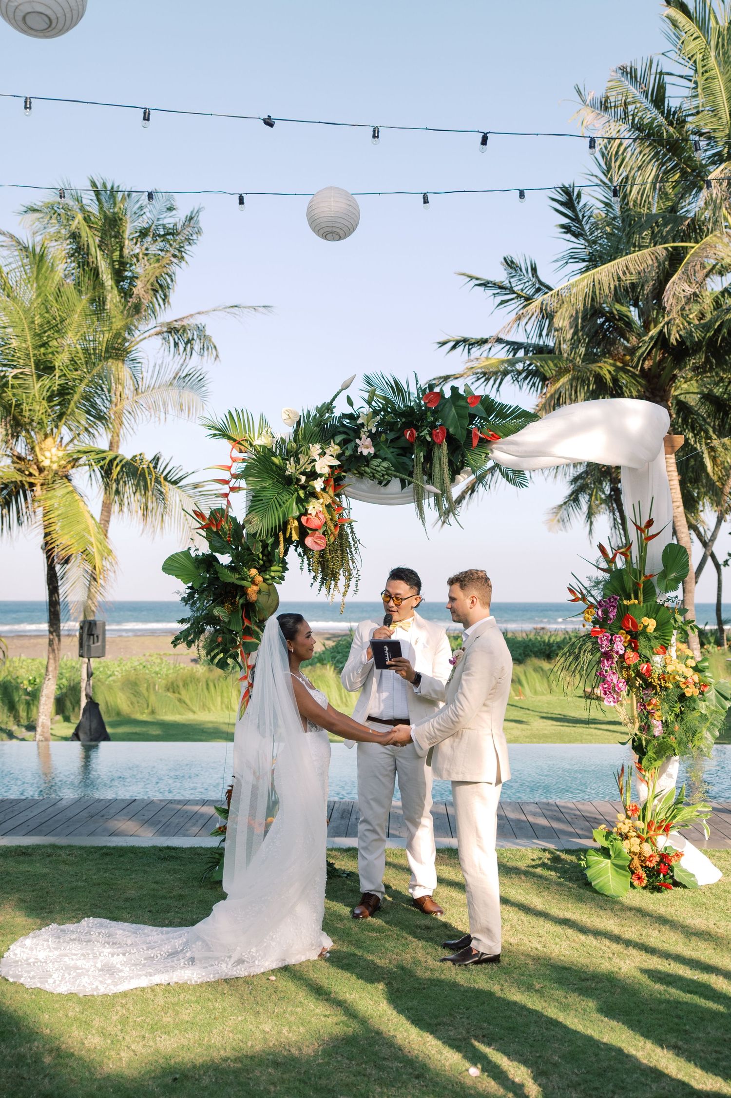 Bride and groom exchange vows under floral arch at beachside tropical wedding ceremony.