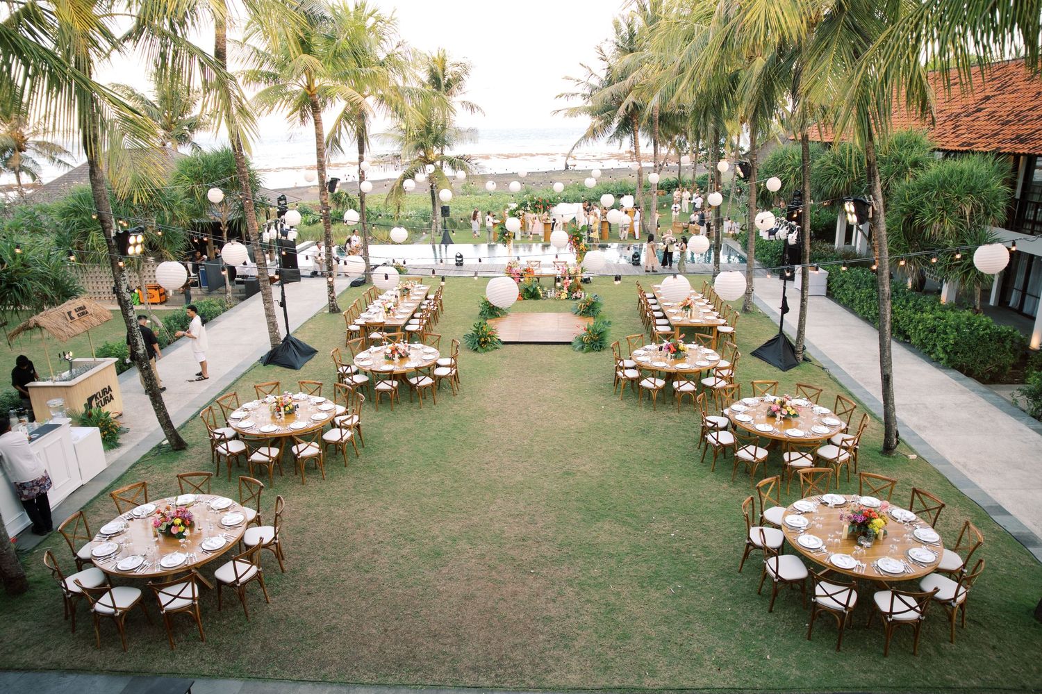Aerial view of a tropical outdoor wedding reception with round tables on a lush green lawn.