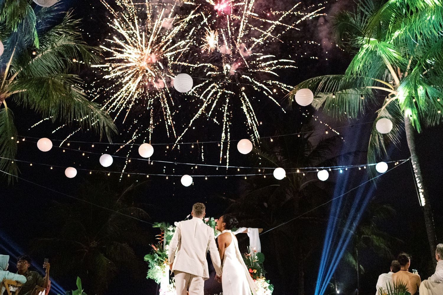Newlyweds embrace on dance floor under glowing lanterns and brilliant fireworks display at tropical night wedding.