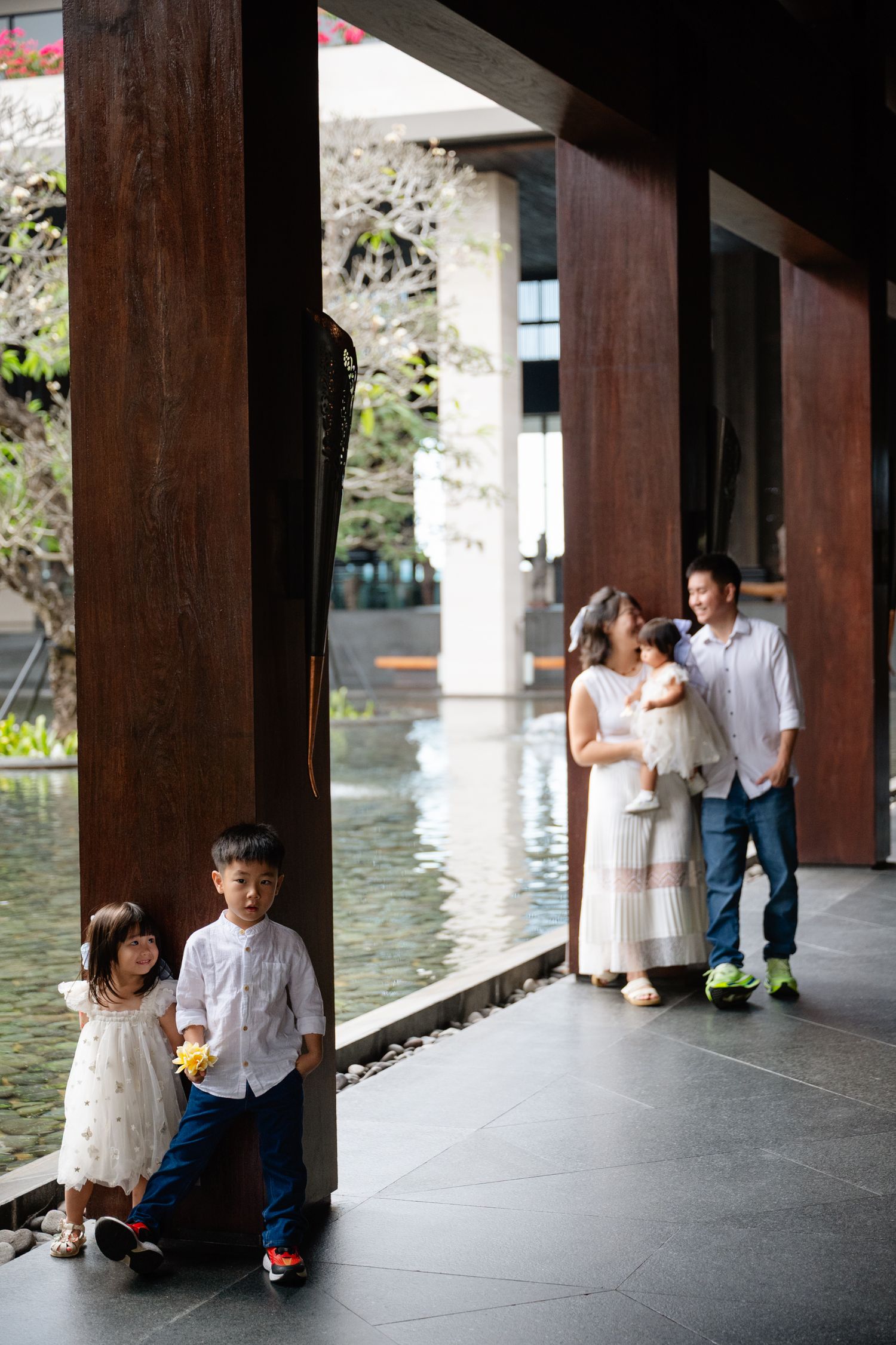 Asian family of four poses in modern courtyard with water feature, two children lean on wooden pillar while parents embrace.