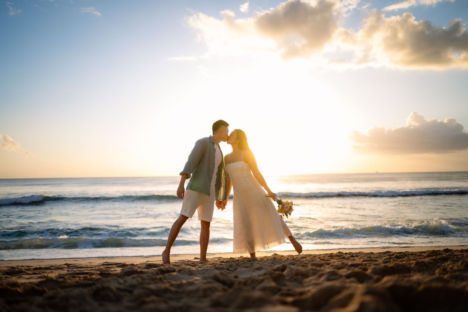 Couple kissing on a sandy beach at sunset with ocean waves and golden sky backdrop.