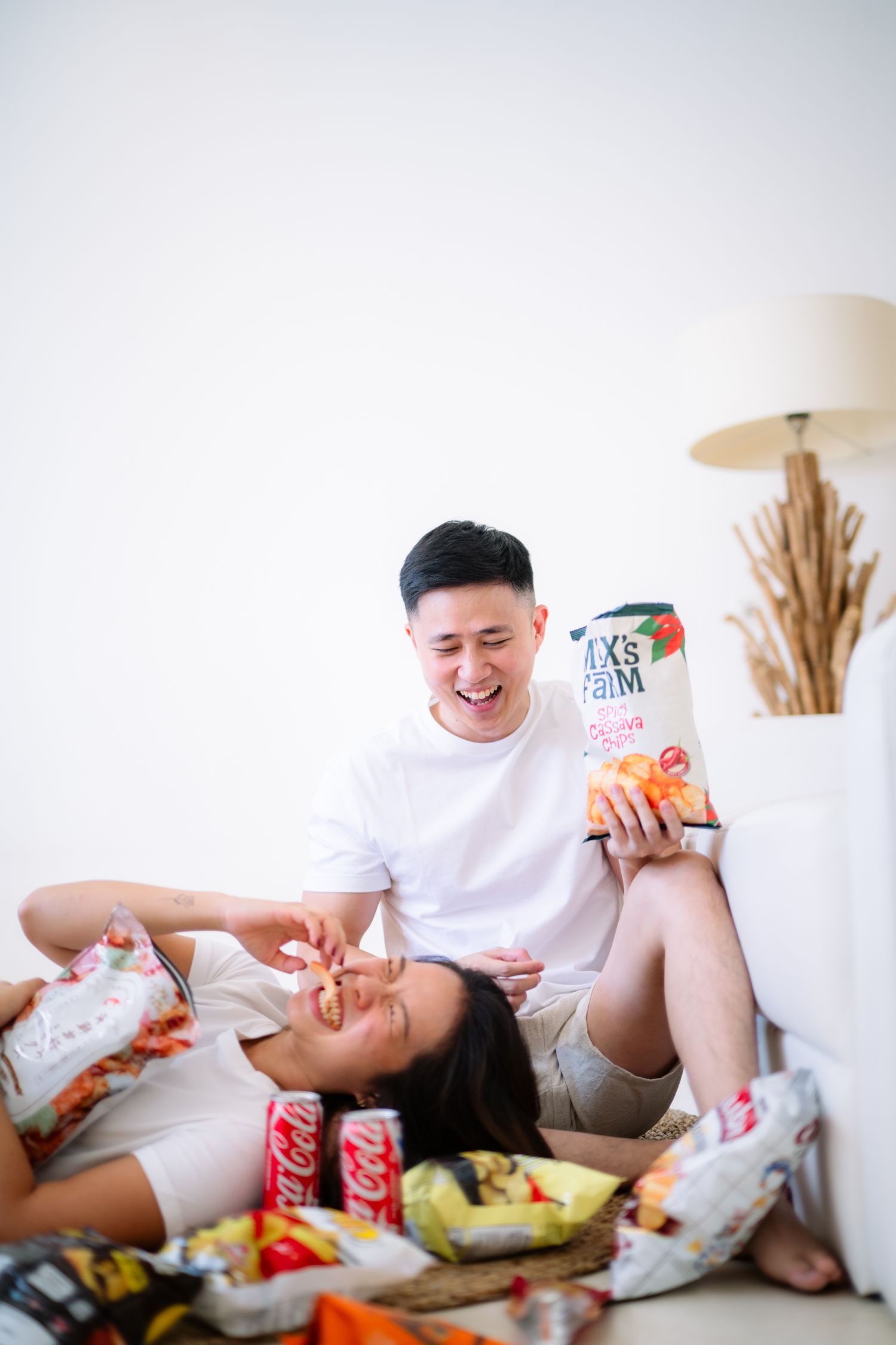Couple enjoying snacks on a white couch, man feeding woman chips while holding a snack bag, surrounded by Coca-Cola cans.