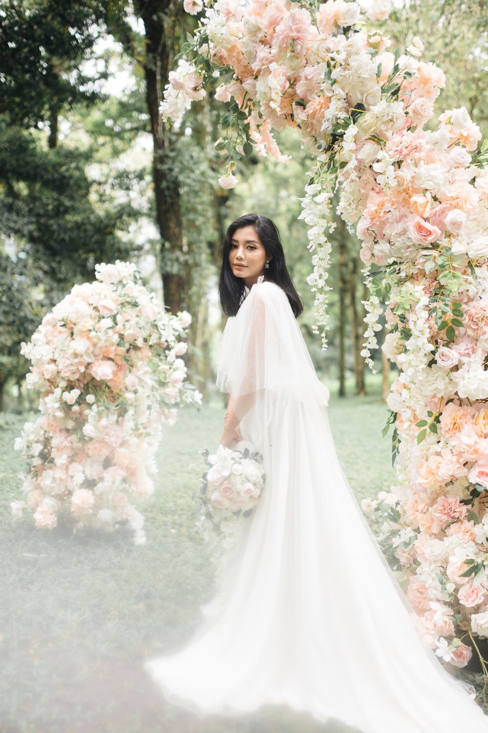 Bride in white cape gown holding bouquet stands beneath floral arch with peach roses in lush garden setting.