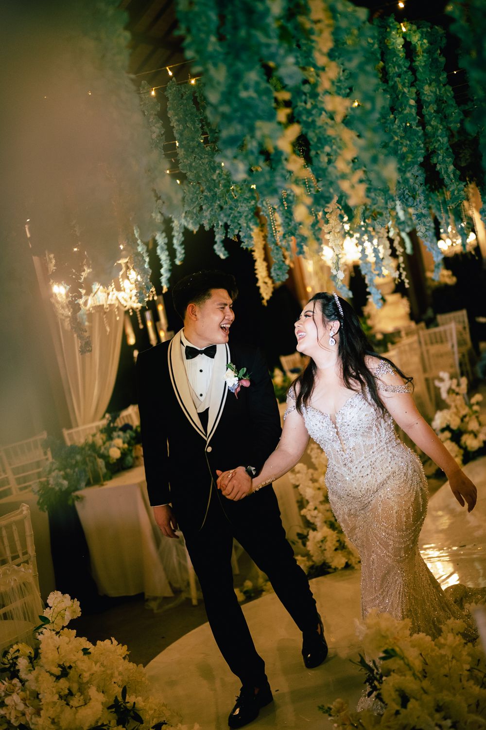 Bride and groom share a joyful moment holding hands under lush floral ceiling decor at elegant wedding reception.