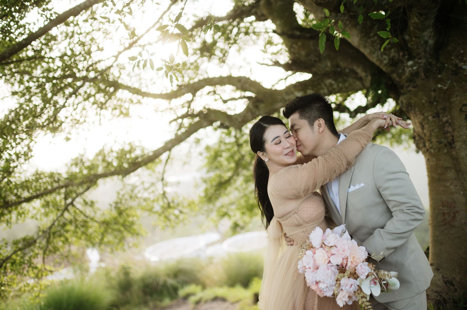 Couple embracing romantically under a large tree, woman holding pink bouquet, outdoor engagement photo session.