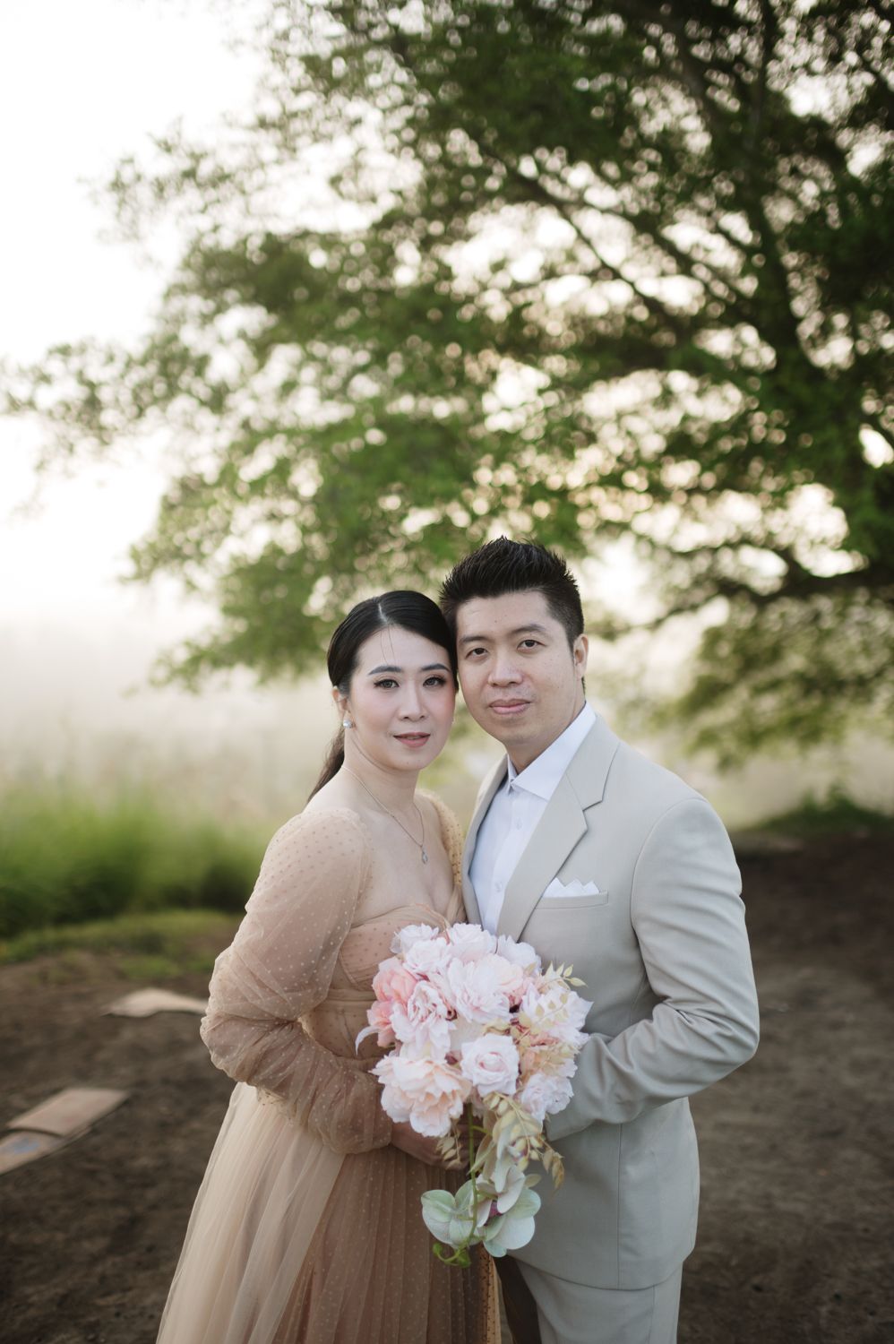 Couple in wedding attire poses outdoors, woman in gold gown holding pink bouquet, man in beige suit, lush green tree background.