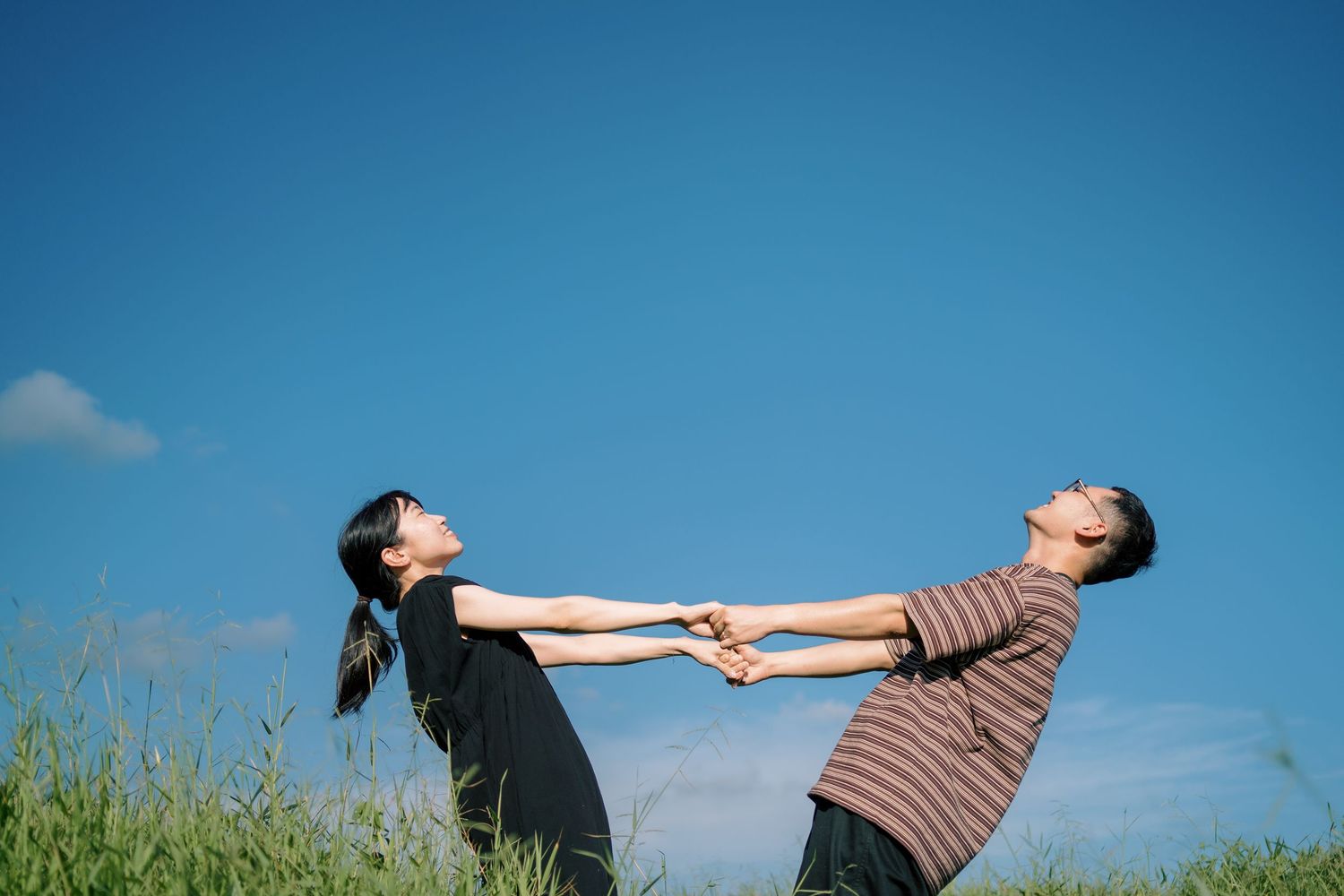 Couple holding hands and leaning back in a green field under a bright blue sky.