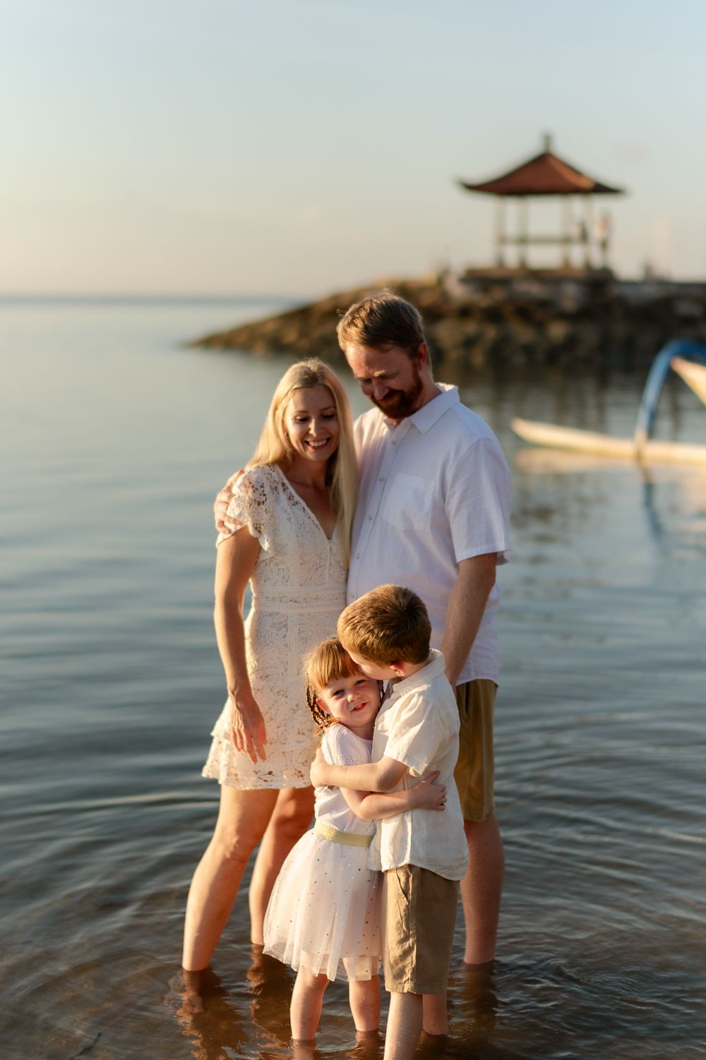Happy family of four standing in shallow beach water with a scenic gazebo at sunset.