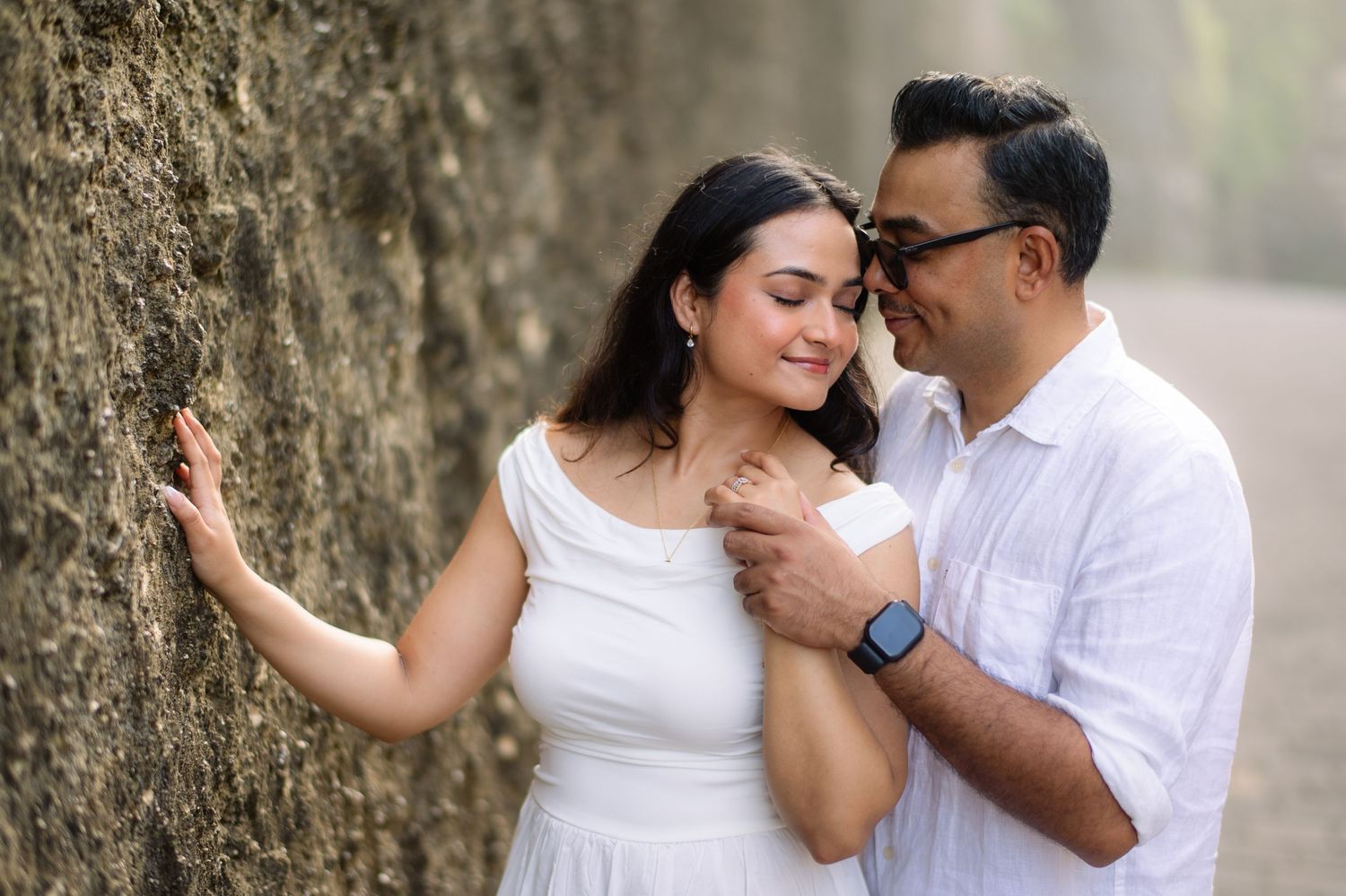 Couple in white outfits sharing a romantic moment against a textured stone wall outdoors.