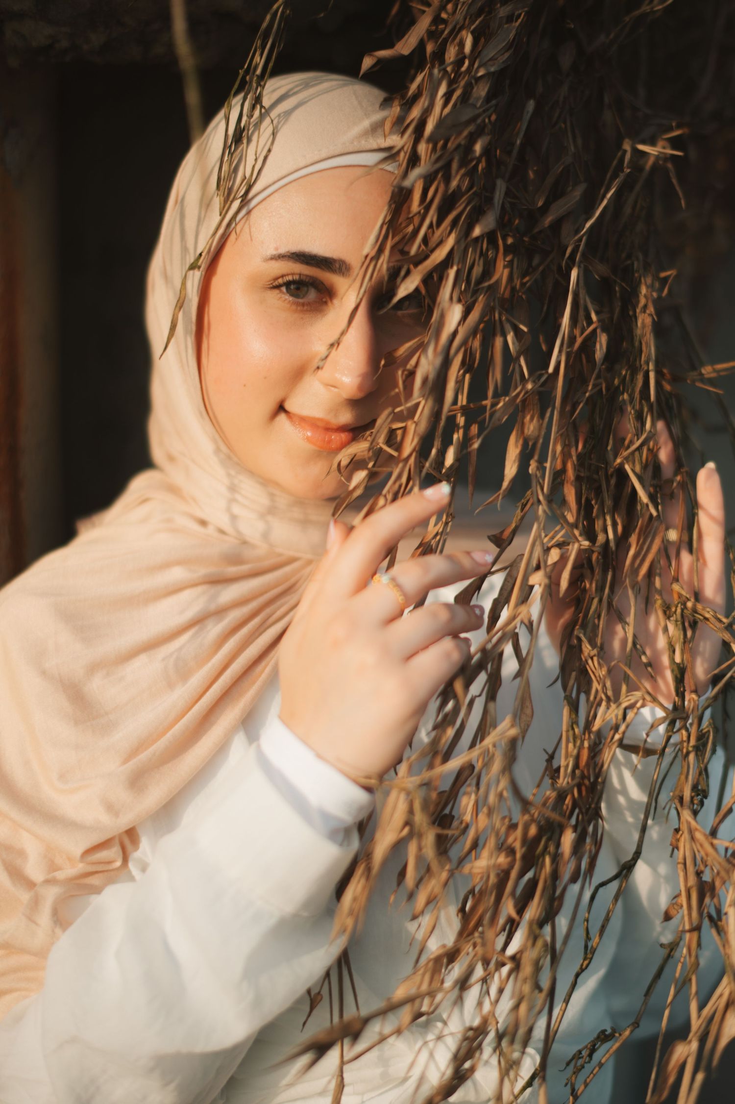 Young woman in beige hijab smiling softly while holding dried branches in warm golden sunlight.