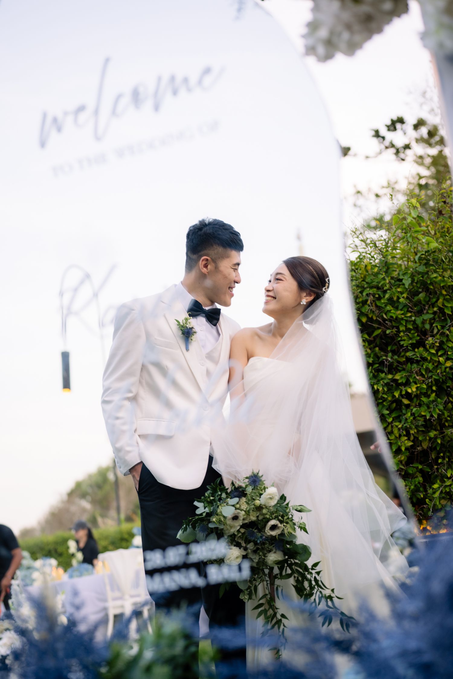 Smiling bride in a veil and groom in a white tuxedo jacket hold a lush bouquet beside a welcome sign at an outdoor garden wedding.