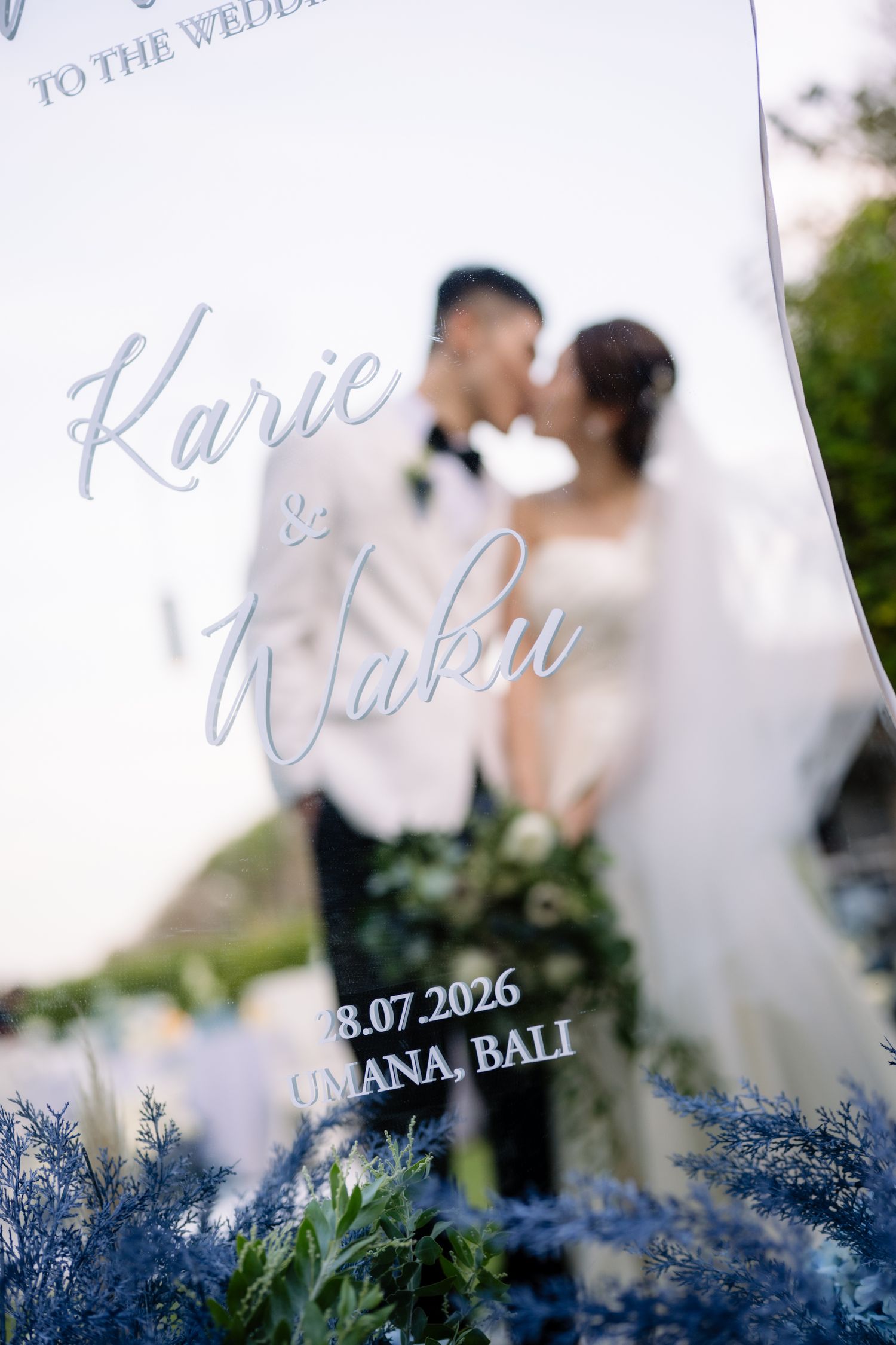 Bride and groom kiss behind an acrylic wedding sign reading Karie & Waku, 18.07.2026, Umana, Bali, with blue floral accents in front.