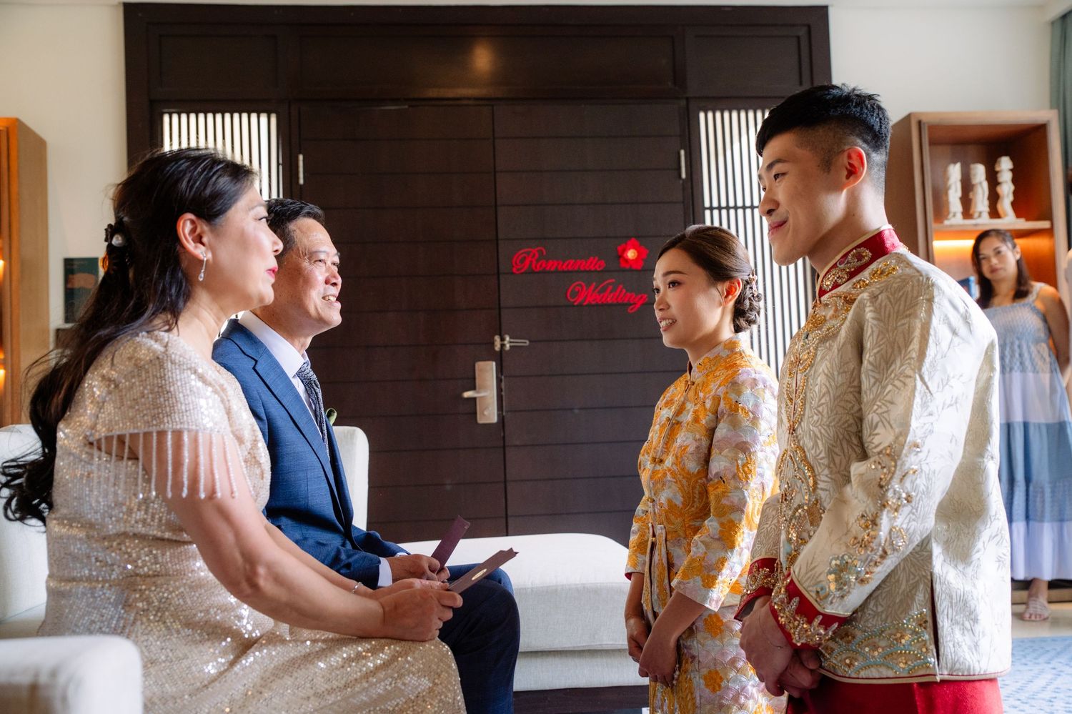 Bride and groom in traditional Chinese wedding attire stand before seated family members during a tea ceremony in a bright living room.