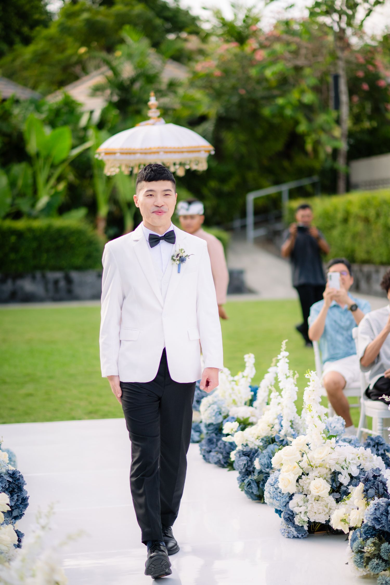 Groom in a white tuxedo jacket and blue bow tie walks down an aisle lined with white and blue flowers at an outdoor garden wedding.